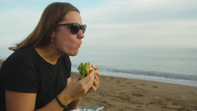 A girl on the seashore, with her mouth wide open, bites off a large piece of burger. Picnic by the sea.