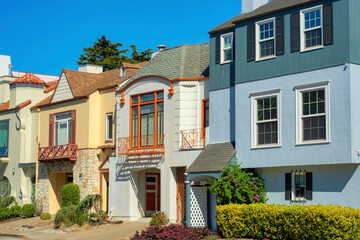 Row of colorful houses beige abd blue and green in midday sun with front yard trees and shrubs with...