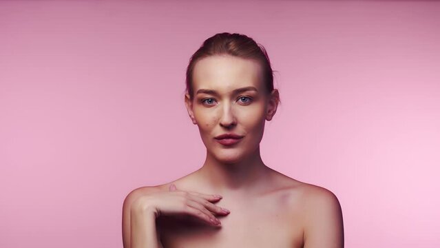 Young beautiful caucasian woman with spectacular makeup slowly touching her own naked collarbones and coquettishly smiling standing on the pink background