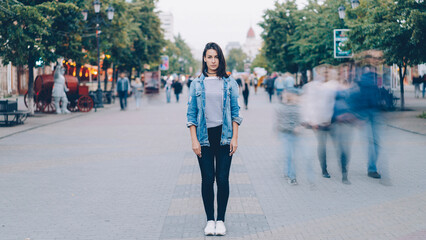 Portrait of pretty girl in trendy clothing looking at camera standing on pedestrian street by herself when busy men and women are moving around in haste. - 550205547