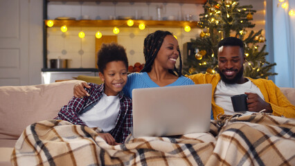 Happy African-American family resting on couch drinking tea and watching movie on laptop on...