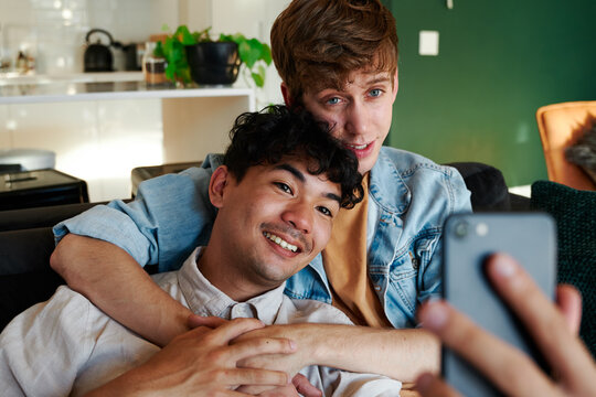 Young gay couple taking selfie with mobile phone while holding hands on sofa at home