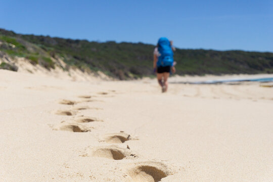 Closeup of footprints on beach while hiker wearing backpack is seen walking along beach with hiking gear in the distance. Sunny day with blue skies along the Cape to Cape track in Western Australia.