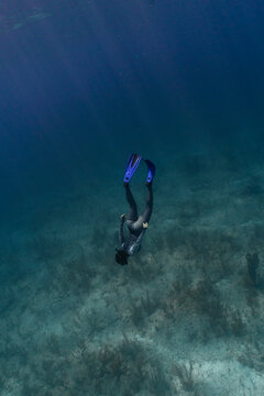 Vertical underwater shot of woman free diving to the bottom of the ocean with sandy grassy sea floor. Women is in lycra top and leggings, sun rays are shining through surface. 