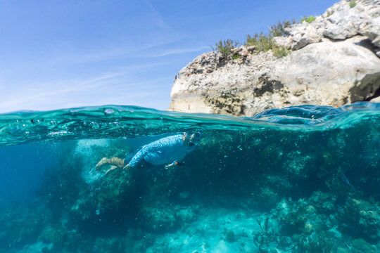 Underwater split shot of man wearing a rashgaurd swiming in clear aqua blue water alongside rocky cliffside in the Bahamas.