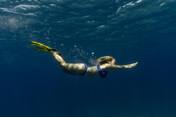 Underwater photo of woman diving into deep blue moody ocean water wearing blue bikini, fins and no mask, long blonde hair flowing behind her. High contrast swimming snorkeling person in Bahamas.
