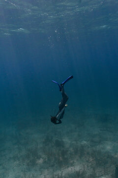 Underwater shot of woman equalizing while freediving down to bottom of shallow ocean in Bahamas with sandy grassy seabed. Sun rays shining through and water is deep blue and moody.