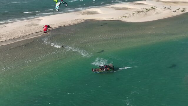 Aerial orbiting shot of kitesurfers performing stunts and jumps amongst boaters and spectators along the coast with a giant sand bar and dunes in tropical waters