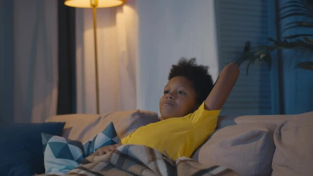 African-American kid sitting alone on sofa holding remote control and watching tv