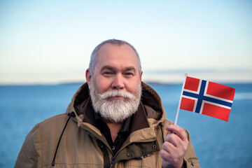 Man holding Norway flag. Portrait of older man with a national Norwegian flag. Visit Norway concept. Older man 50 55 60 years old with gray beard outdoors travelling. Travel to Norway concept.  - 550219737
