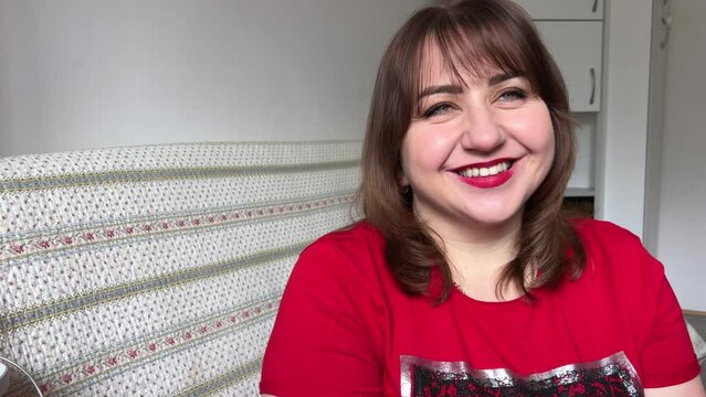 a plump woman, curvy and appetizing, sits in front of the camera and smiles, she has red lipstick, a red jacket and brown dark hair
