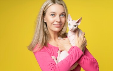Portrait of blond woman in red with cat in hand. Attractive charming Woman holding Canadian sphynx cat in hands isolated on yellow background. Woman and Cat with big green eyes looking at camera.