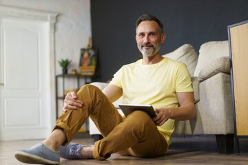 Fototapeta premium portrait of adult man dressed in yellow t-shirt sitting on the floor at home, leaning back on sofa with tablet pc in his hands. Looking at camera and smiling
