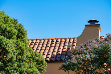 Suburban roof facade on house or home with adobe red tiles and visible chimney with stucco beige paint and metal top in late sun