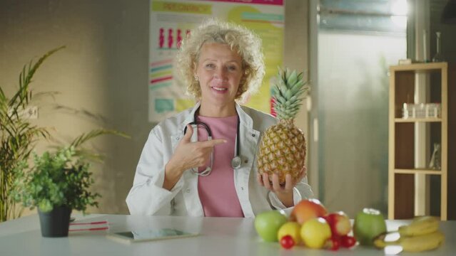 A mature female doctor or nutritionist sitting at the table with different fruits and pointing at pineapple
