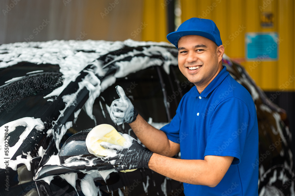 Wall mural Man worker washing car service with foam and sponge. Car wash cleaning wipe station. Employees clean a vehicle professionally.