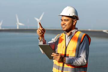 Engineer India man working with tablet at windmill farm hydroelectric Generating electricity clean energy. Wind turbine farm and water power generator by green energy. Engineer control electric power
