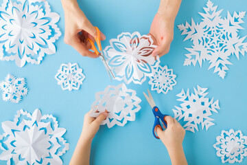 Adult mother and child hands cutting different white snowflake shapes from paper on light blue table background. Pastel color. Making decoration elements for winter festive. Closeup. Top down view.