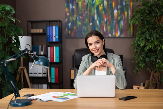 Photo of delighted young woman wearing headphones looking at camera smiling sitting at desk in front of computer laptop working studing process from home.