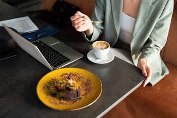 Close up shot above woman's hands holding tea spoon with coffee foam.Laptop cappuccino and dessert on table. Woman having break after work relaxing at restaurant caffe.