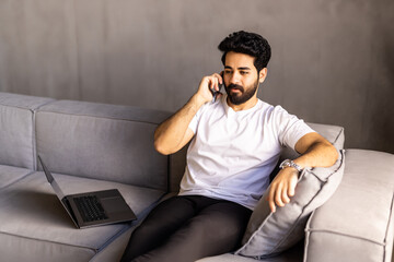 Fototapeta premium Cheerful arab man with long hair and glasses relaxing on couch at home, having phone talk with friends, looking at copy space and smiling, wearing casual outfit, resting at weekend