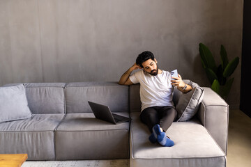 Naklejka premium Cool Gadget And Application. Portrait of young smiling Arab man holding mobile phone, typing sms message, sitting on the couch in living room.