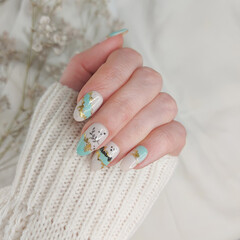 Beautiful woman's hands with nude cozy manicure design in white, milky, turquoise colors, decorated with gold. Long fingers holding a knitted winter sweater.
