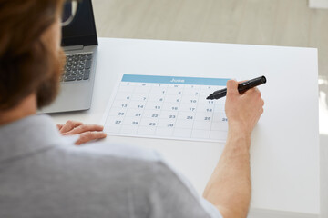 Busy man organizes his working schedule. Businessman sitting at his office desk with a laptop and marking dates on his business calendar. View over the shoulder. Cropped shot. Planning time concept