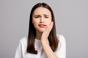 Crying tired sad woman suffers from painful toothache dental illness pointing on teeth standing on grey background in studio isolated wearing basic white t-shirt.