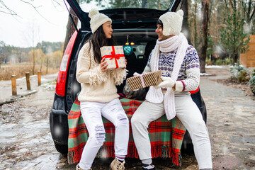 Beautiful young couple giving presents and looking each other sitting in the trunk in winter holidays.