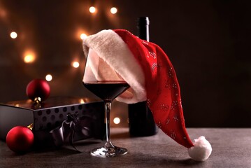 Glass of red wine with Santa Claus hat, black gift box and Christmas decorations on table. Christmas lights in the black background. Copy space for text.