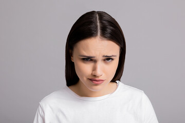 Sad young desperate woman in white casual t-shirt showing depressive emotions standing alone on grey background.