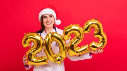 A portrait of a joyful nurse in a white coat and a Santa Claus hat holds the figures of the new...