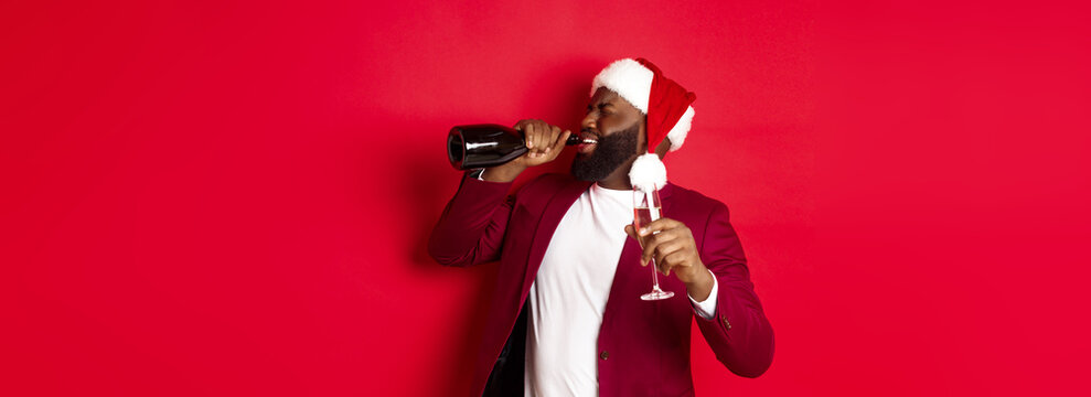 Christmas, party and holidays concept. Image of young Black man in santa hat drinking champagne from bottle, getting drunk on New Year celebration, standing over red background
