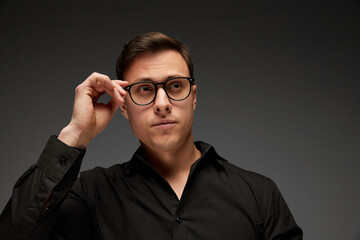 Serious young man, manager in black business classic style shirt putting eyewear on isolated over grey background. Fashion look of businessman. Office style