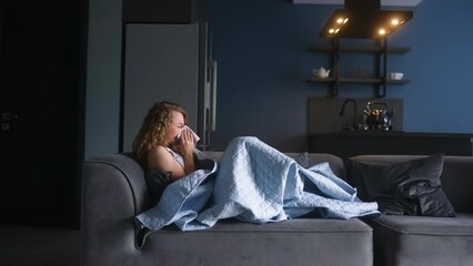 Woman sick with flu coughing and blowing her nose under blanket on couch at home. Young caucasian female suffering from coronavirus Covid19 while isolating. Ill girl laying on a sofa struggling cold