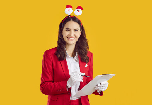 Smiling happy brunette woman wearing in Santa style with Xmas hair band in Santa shape making notes in clipboard on yellow background looking at camera. New Year, Christmas, Xmas social survey.