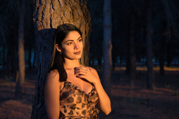 Fototapeta premium South American woman, young and beautiful, brunette, in leopard dress, leaning on the trunk of a tree, in the golden hour. Concept beauty, fashion, autumn, golden hour, melancholy.