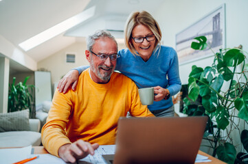 Senior middle aged happy couple using laptop together at home - obrazy, fototapety, plakaty