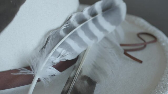 Close-up view of white and gray striped feathers in white hat with leather wrap. Custom beadwork brim. Two-spirit wedding outfit. Accessories.