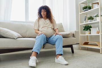 Fototapeta premium Pregnant woman sitting on the couch at home and wearing pregnancy-friendly shoes with a problem due to the stomach, headache, pregnancy and motherhood difficulties, severe fatigue, headache