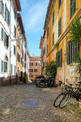 A narrow Italian street, with bicycles, motorcycles, and potted plants on the right. Ahead there is a building with yellow stucco.