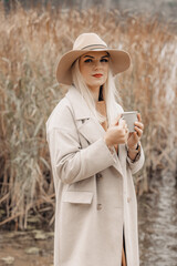 A young woman in a coat and hat enjoy nature and a cup of hot tea on the shore of a lake on an autumn day