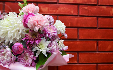Opposite the red brick wall is a beautiful bouquet of flowers. A bouquet of peony buds, pink carnations, white chrysanthemums are organically combined into a single composition. Postcard for the holid