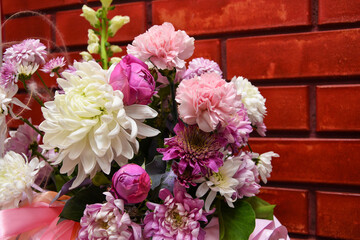 Fototapeta na wymiar Opposite the red brick wall is a beautiful bouquet of flowers. A bouquet of peony buds, pink carnations, white chrysanthemums are organically combined into a single composition. Postcard for the holid