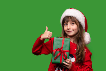 Smiling little girl holds a gift on a green isolated background. A child in a Santa Claus hat and a warm sweater holds a gift box.