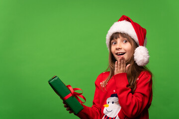 Cute smiling little Santa girl in a hat holds a New Year's gift in her hands and looks happily at the camera. Time to give gifts. Merry Christmas and Happy New Year. Isolated on green background