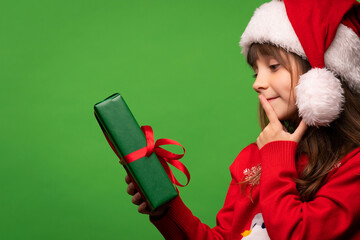 A cute little Santa girl in a hat holds a New Year's gift in her hands, looks at it dreamily and tries to guess the surprise in the box. Merry Christmas and Happy New Year. Isolated on green