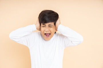 Frustrated little Indian boy child screaming with shut ears standing over beige studio background. disturbed kid.