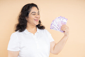 Happy senior Indian woman holding money of 100 rupee notes isolated on beige background, Asian mature female with lot of cash in hand.